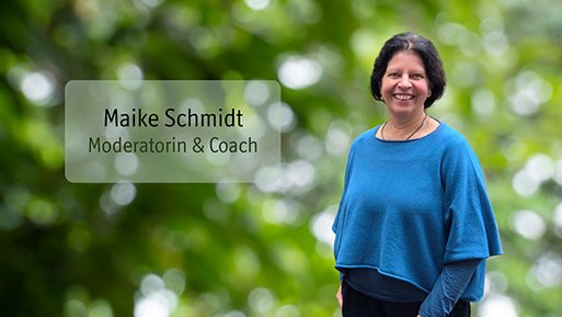 Maike Schmidt Frau mit dunklen Haaren und blauem Pullover steht vor unscharf grünem Hintergrund und lächelt.
