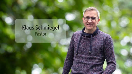 Klaus Schmiegel Mann mit Brille und Kapuzenpullover steht vor unscharf grünem Hintergrund mit Textfeld 'Klaus Schmiegel Pastor'.