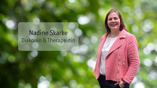 Nadine Skarke Frau mit rosa Jacke und weißem Oberteil steht vor unscharf grünem Hintergrund und lächelt.