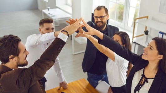 Mehrere Personen stehen um einen Tisch und legen ihre Hände in der Mitte übereinander. Sie tragen Businesskleidung und wirken konzentriert. Im Hintergrund sind große Fenster und helle Büroräume zu sehen.