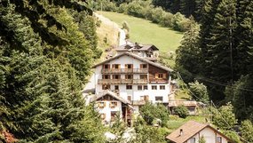 Sommer-Singwoche Südtirol Mehrstöckiges Haus mit Holzbalkonen in bewaldeter Berglandschaft mit Wiesen und Nadelbäumen.