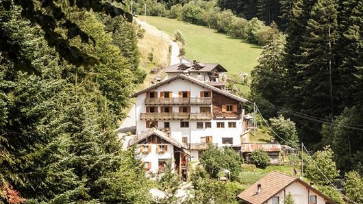 Sommer-Singwoche Südtirol Mehrstöckiges Haus mit Holzbalkonen in bewaldeter Berglandschaft mit Wiesen und Nadelbäumen.