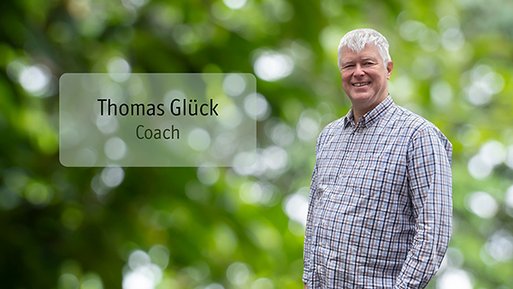 Thomas Glück Mann mit kariertem Hemd steht vor unscharf grünem Hintergrund und lächelt.