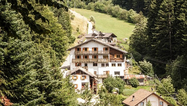 Sommer-Singwoche Südtirol Mehrstöckiges Haus mit Holzbalkonen in bewaldeter Berglandschaft mit Wiesen und Nadelbäumen.