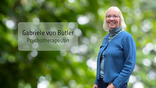 Gabriele von Butler Lächelnde Frau mit Brille und blauem Cardigan vor unscharf grünem Hintergrund.